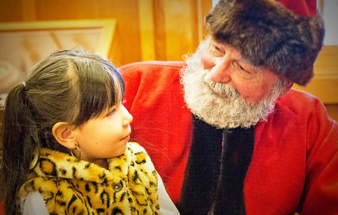 little girl and santa look at each other as girl sits on his lap.