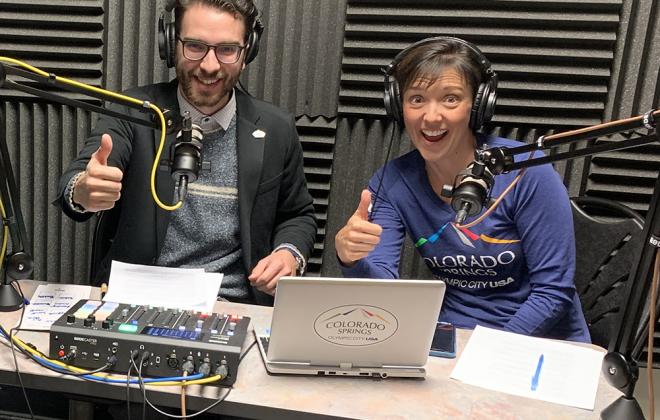 Jen and Ted give thumbs up from the audio booth. Gray sound proof materials on the way behind them, studio microphones and sound board in front.