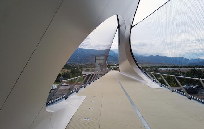 looking down the path of the bridge. on the left, a curved wall that loops overhead and to the other side. there are large curved gaps in the wall on each side. The effect is a wave that moves from one side of the bridge to the other.